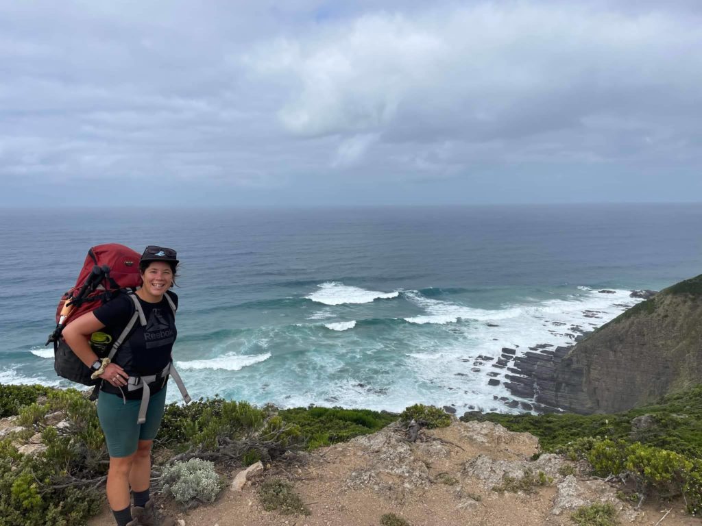 A rocky outcrop with small shrubs in the foreground. A woman with a red hiking pack is standing posing to the left of the photo. In the background you can see the ocean with rolling waves towards a rocky cliff face. The sky is overcast and grey.