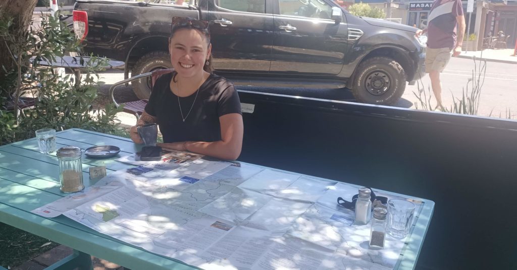 Great Ocean Walk Map is spread out on outdoor cafe table with a coffee and a woman sitting at the table smiling.