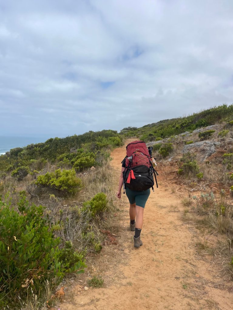 A sandy trail with a woman hiking with a red pack on. The sky is full of clouds you can barely see any blue sky.