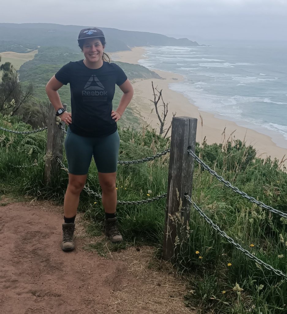 A woman standing proudly beside a chain fence on a cliff edge facing the camera. Behind the cliff is a beach and rolling hills. The ocean has waves and the sky is so grey you can't differentiate the clouds.