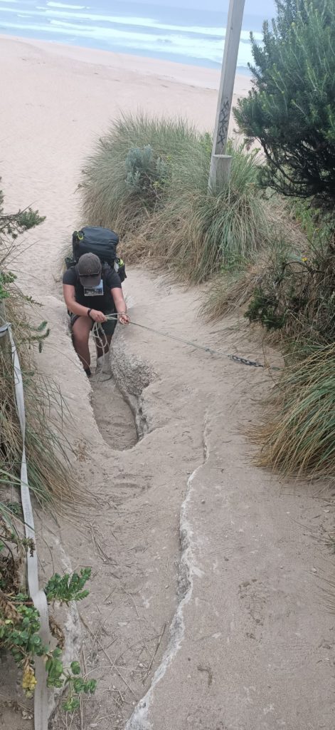 A woman has a blue backpack on, is facing the camera and pulling on a rope to help her walk up a rock exit from the sandy beach.