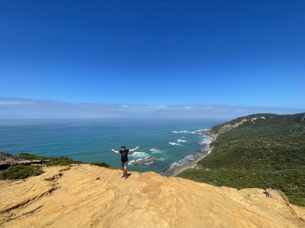 A massive flat rock on the top of a cliff, looking down a the rugged coast line. In the background the ocean and a blue sky, including the hilly coastline covered in coastal brush. 