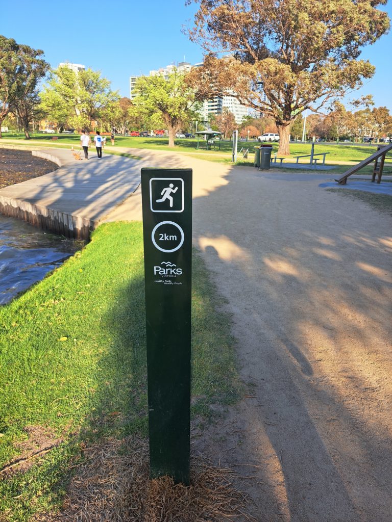 A view of the sandy trail with a green km marker centre picture. There is a workout zone in the background and two people in white tshirts walking along the trail in the distance.