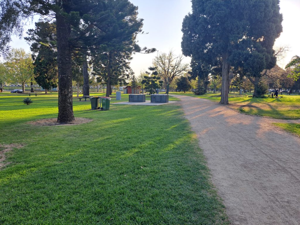 There is a sandy path with bbqs to the left of it and various trees in a picnic area in the background of the picture is the red toilet block.