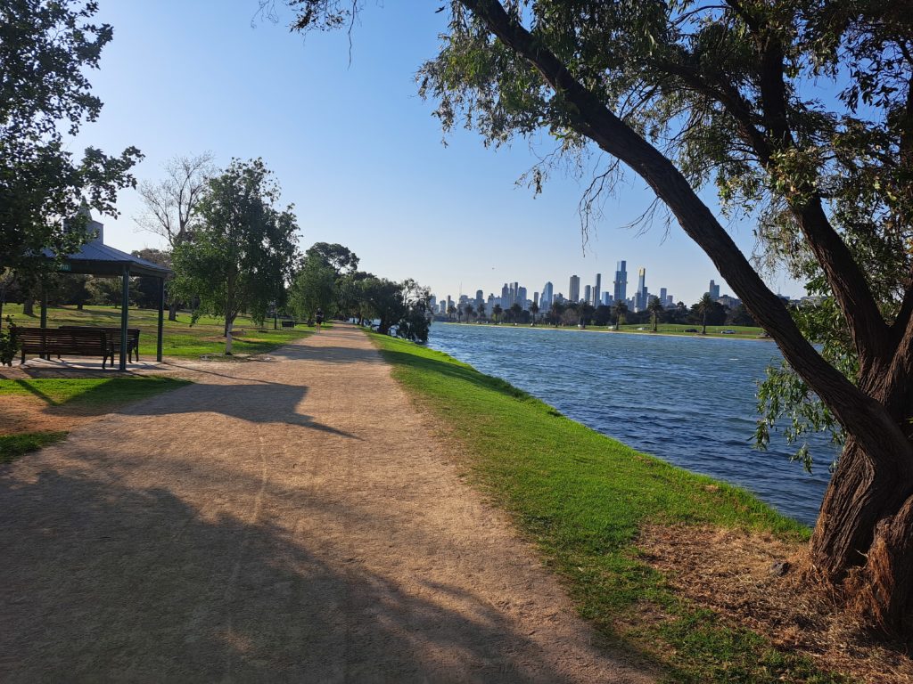 Lake view of the city skyline to the left is a shelter and trees along the pathway. The sky is blue and clear.