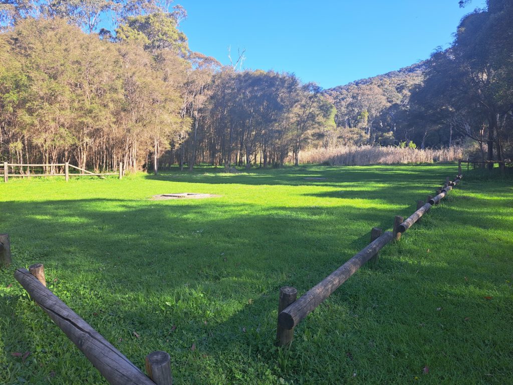 A grassy campsite surrounded by a low lying wooden barrier and a campfire in the centre.