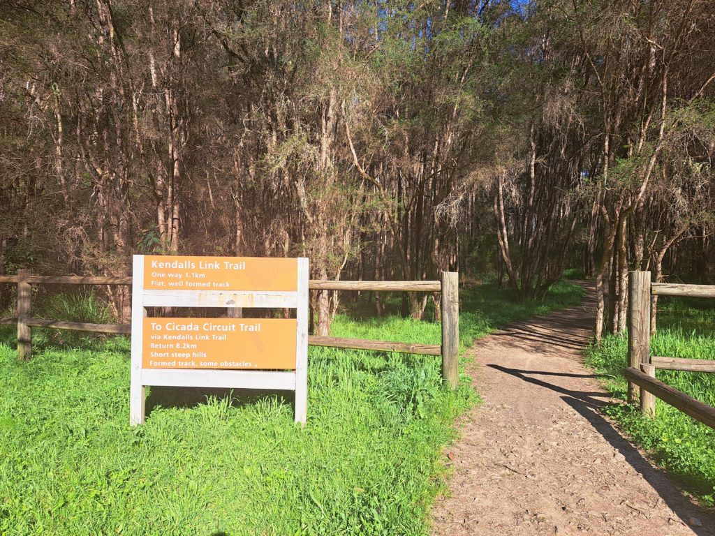 A view of the trail passing through a fence line with a sign explaining the hiking options, there are skinny teatrees in the background.