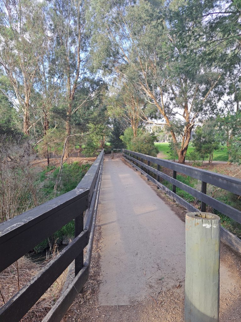 Concrete sturdy bridge with blue painted wooden handrails across the creek. There are many trees in the background.